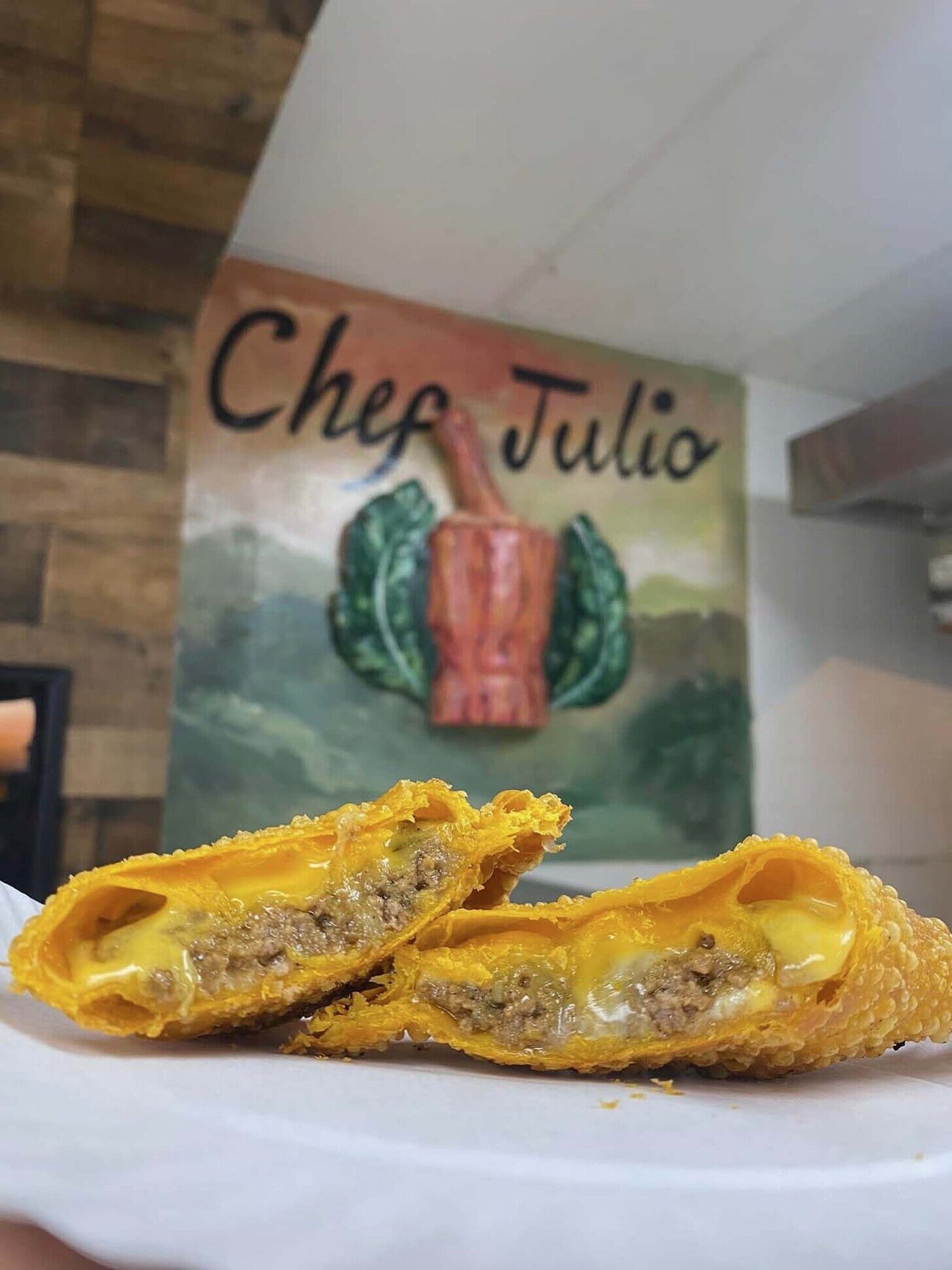 Golden corn empanada split open in front of the Chef Julio mural, showing melted cheese filling